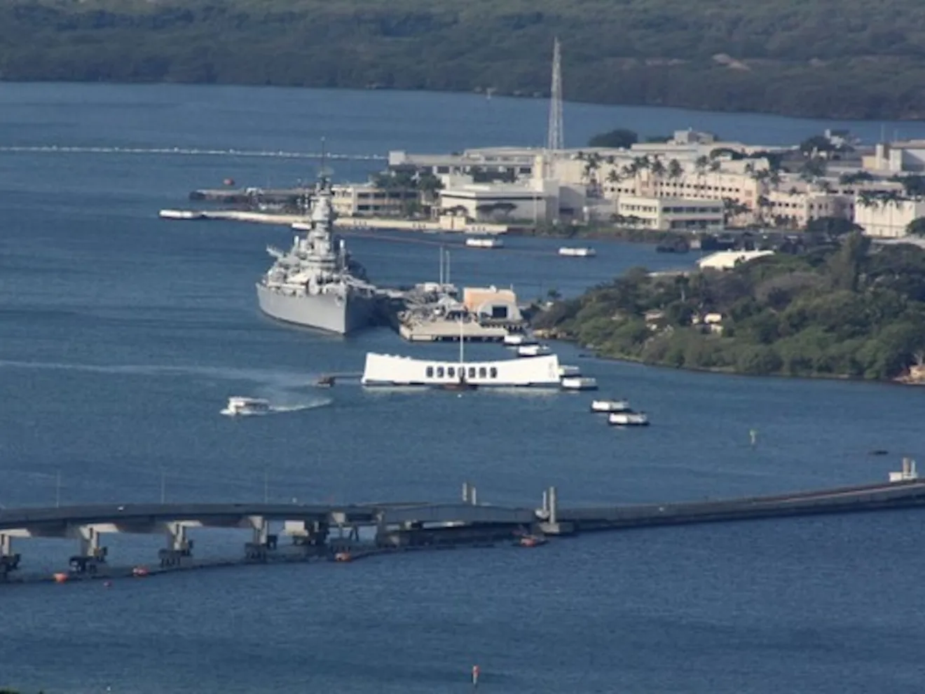 Aerial view of a naval ship and memorial on a coastline with bridge and buildings in the background.